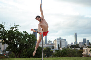 Muscular male pole dancer, holding a pose on a pole set outdoors. Tel Aviv buildings in the far...