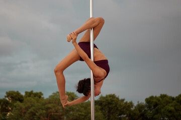 Young Middle Eastern pole dancer, holding a pose on a pole set in an urban park.