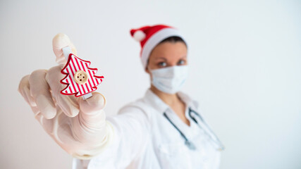 Female medical worker with protective equipment and Santa hat holding a small Christmas tree in his hand. Doctor during the Christmas and New Year holidays.