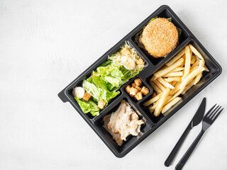 Lunch box with caesar salad, burger and fries in black plastic package