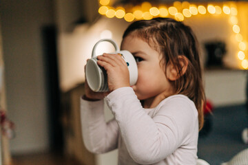 Caucasian toddler girl drinking cocoa from mug at home. Christmas bokeh lights