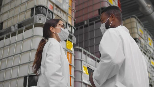 African and caucasian inspectors in safety mask and lab coat standing in chemical factory warehouse