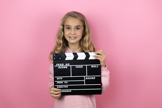Young beautiful child girl standing over isolated pink background holding clapperboard very happy