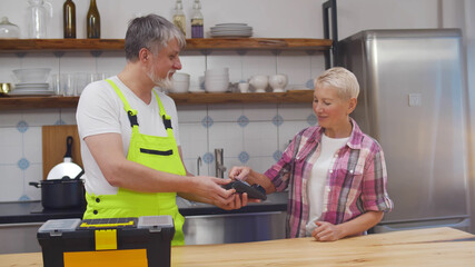 Senior repairman in overall holding wireless terminal for client to pay for service.