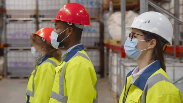 Side view diverse team of industrial workers in helmet and safety mask walking in large warehouse