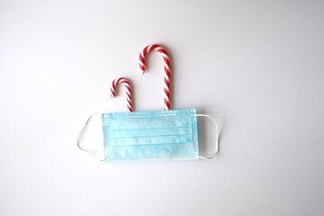 Flat lay. Christmas, new year and coronavirus theme. Two red and white candy canes are covered with a medical mask. Located in the center. White background.