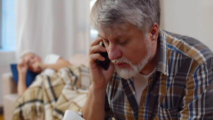 Worried aged husband holding infrared thermometer calling doctor with sick wife lying on background