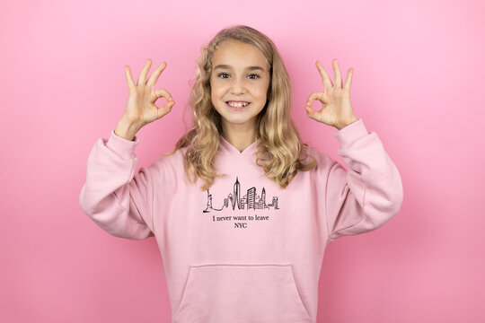 Young beautiful child girl standing over isolated pink background doing ok sign with fingers and smiling, excellent symbol