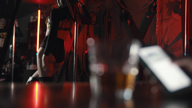 Close up of man sitting at bar counter and using smartphone with stripper dancing on background
