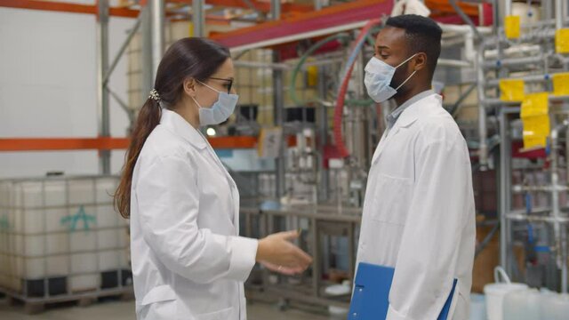 Interracial warehouse inspectors in safety mask and lab coat do elbow bump greeting