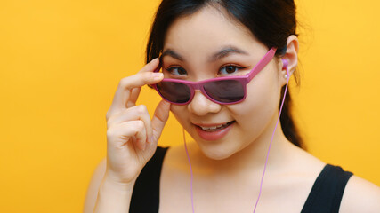 Fototapeta premium Portrait of young asian woman listening to the music on the headphones and looking over her pink sunglasess. Isolated on orange background. High quality photo