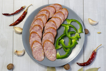 Appetizing sausage sliced on a grey plate on a light wooden background with green bell pepper, chilli, close up