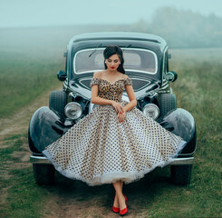 Young beautiful sexy woman in pin-up style clothes posing near black retro car. Polka dot white dress, vintage hairstyle, red high heels. Background road green nature fog. Girl fashion model driver - obrazy, fototapety, plakaty