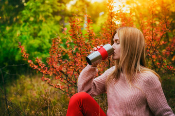 Female drinks hot coffee from a thermos in an autumn park.