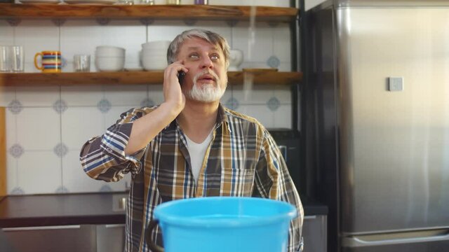 Worried aged man calling plumber while water falling into bucket from ceiling at home
