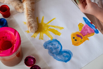A little girl paints the sun and her mother with watercolors, a ginger cat lies next to the table. Child's drawing with sun and mom. Distance learning, close up