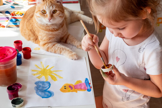 A little girl paints the sun and her mother with watercolors, a ginger cat lies next to the table. Children's creativity. Distance learning