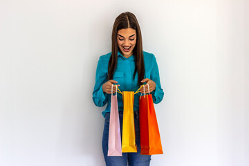 Caucasian woman holding colorful shopping bags, posing on white background. Girl with big smile enjoying shopping sales. Shocked young woman in trendy outfit looking at purchases inside paper bag.