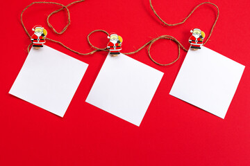 New year's holiday background. White sheets for notes on wooden clothespins with figures of Santa Claus, on a jute rope, on a red background. The view from the top. Greeting card for Christmas.