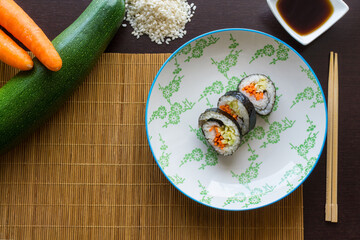 Overhead view of vegan sushi plate on traditional mat. Carrot, zucchini, white rice, soy sauce and chopsticks on wooden background.