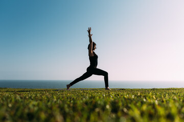 Mujer adulta delgada estira sus brazos al cielo practicando yoga en la tarde en exteriores. - obrazy, fototapety, plakaty