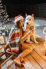 Portrait of a woman in christmas hat and plaid with her cute dog celebrating a New Year holidays at home, feeding dog with gingerbread cookies and making selfie photo