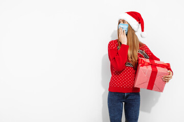 Shocked young woman in Christmas Santa Claus hat, wearing a medical protective mask on her face, with a gift box on a white background