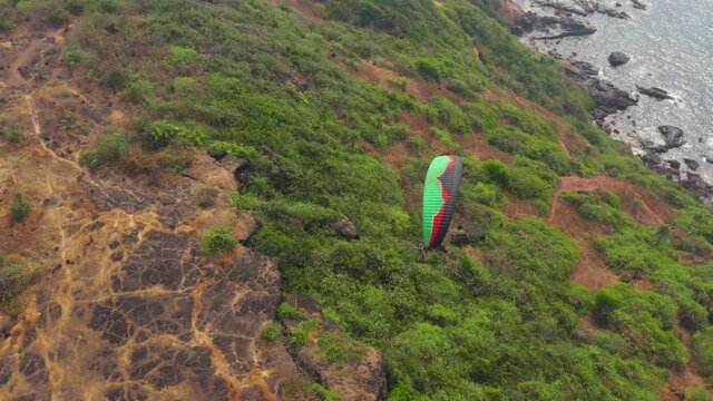 Person flies on a green and black paraglider over a hill and trees to the sea waves near the rocks