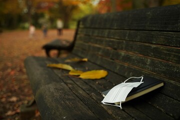 White surgical mask and a book are on a bench in a park in autumn. Mask is for allergy to pollen and also for coronavirus,COVID-19.