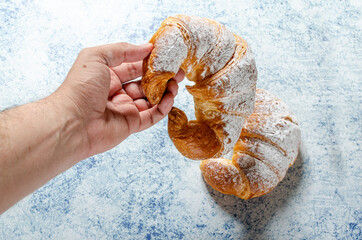 Hand holding a freshly baked croissant with icing sugar on a textured blue background