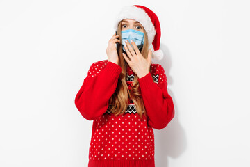 Shocked Christmas woman, wearing a Santa Claus hat and a medical mask on her face, talking on the phone, holding a smartphone, , on a white background