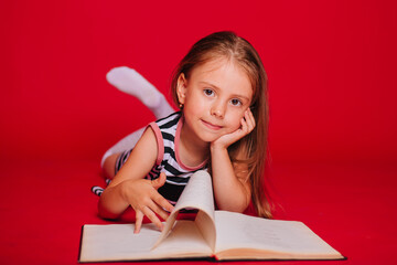 A little girl with blond hair in a striped dress is reading a book. Child on a red background. Learn to read. Preparation for school. Self-isolation lessons