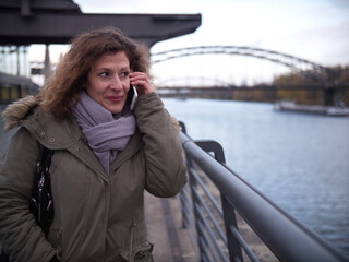 Fototapeta premium Woman stands next to a the river Main and is on the phone. She smiles and seems happy. City Frankfurt am Main during autumn.