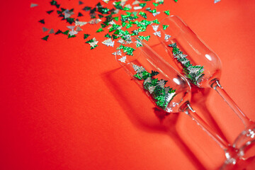 Two champagne glasses with splashing confetti in the form of Christmas trees on a red background. New Year concept