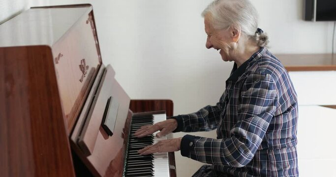 An old lady with gray hair plays the piano. A grandmother with deep wrinkled fingers presses the white keys of a musical instrument.