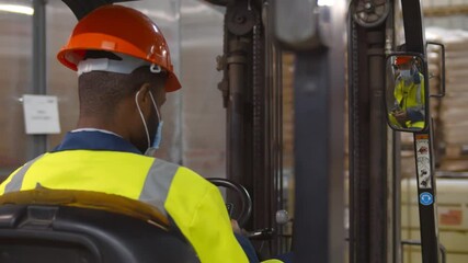 African warehouse worker wearing helmet and safety mask sitting in forklift using smartphone