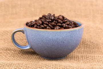 Blue ceramic cup filled with brown roasted coffee beans isolated over burlap