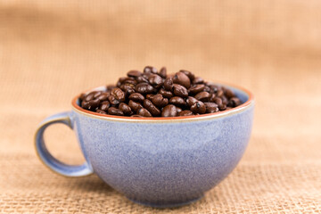 Cup filled with brown roasted coffee beans isolated over burlap very blurry background