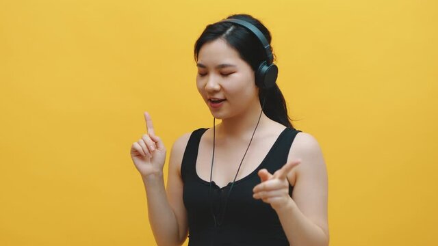 Happy young asian woman with headset listening to the music and dancing. Isolated on yellow background. High quality 4k footage