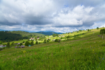 Fototapeta premium Rural mountain landscape, village and geen meadows, mountains on the horizon, dark sky above.