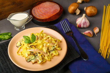 pasta with ham and mushrooms in a creamy sauce on a black board and a blue napkin with a fork near the ingredients.
