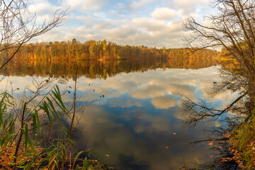 The risen sun illuminates the autumn forest. The cloudy sky is reflected in the crystal blue water of the lake.