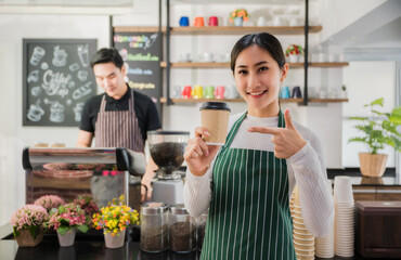 Beautiful happy asian young barista woman in apron holding cup coffee with serving to customer in cafe. Portrait of girl barista standing front counter smiling and point coffee glass in coffee shop.