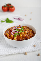 Chole masala or chana local indian street food made of cooked chickpeas, tomatoes and cumin decorated with red onion rings and parsley served on textile on white wooden background at kitchen. Vertical