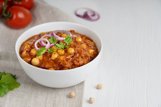 Chole masala or chana indian street food made of chickpeas, tomatoes and cumin decorated with red onion rings and parsley served in bowl on textile on white wooden background. Image with copy space