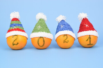 Four tangerines in knitted Christmas caps on a blue background. The tangerines bear the number 2021, one digit on each.