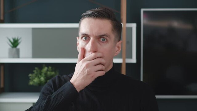Surprised and shocked young business man in black sweater is opening her mouth at a loss and looking to the camera on modern interior background. 4k