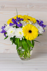 A bouquet of bright flowers in a glass jug on a light wooden background. Still life in a rustic style. Yellow gerbera, blue iris and white chrysanthemum in a vase. Festive postcard for mother's day.