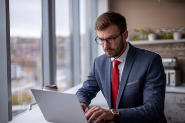 Young businessman typing on a laptop in an office near a window.