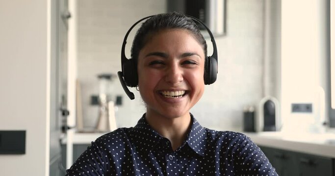 Close up head shot web camera view overjoyed young indian mixed race woman in headphones with microphone laughing at joke, having funny online video call conversation with friends or colleagues.